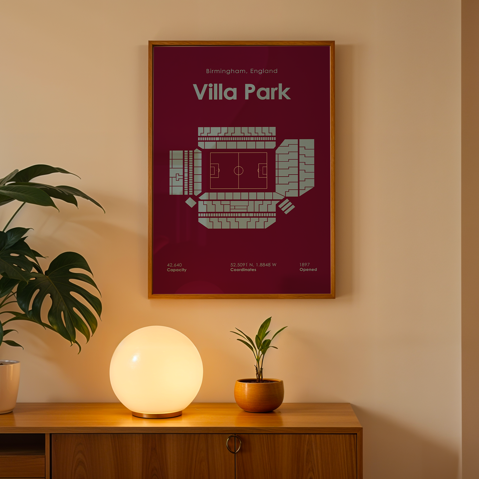 Framed poster of Villa Park stadium on a wall with a lamp and plant below.