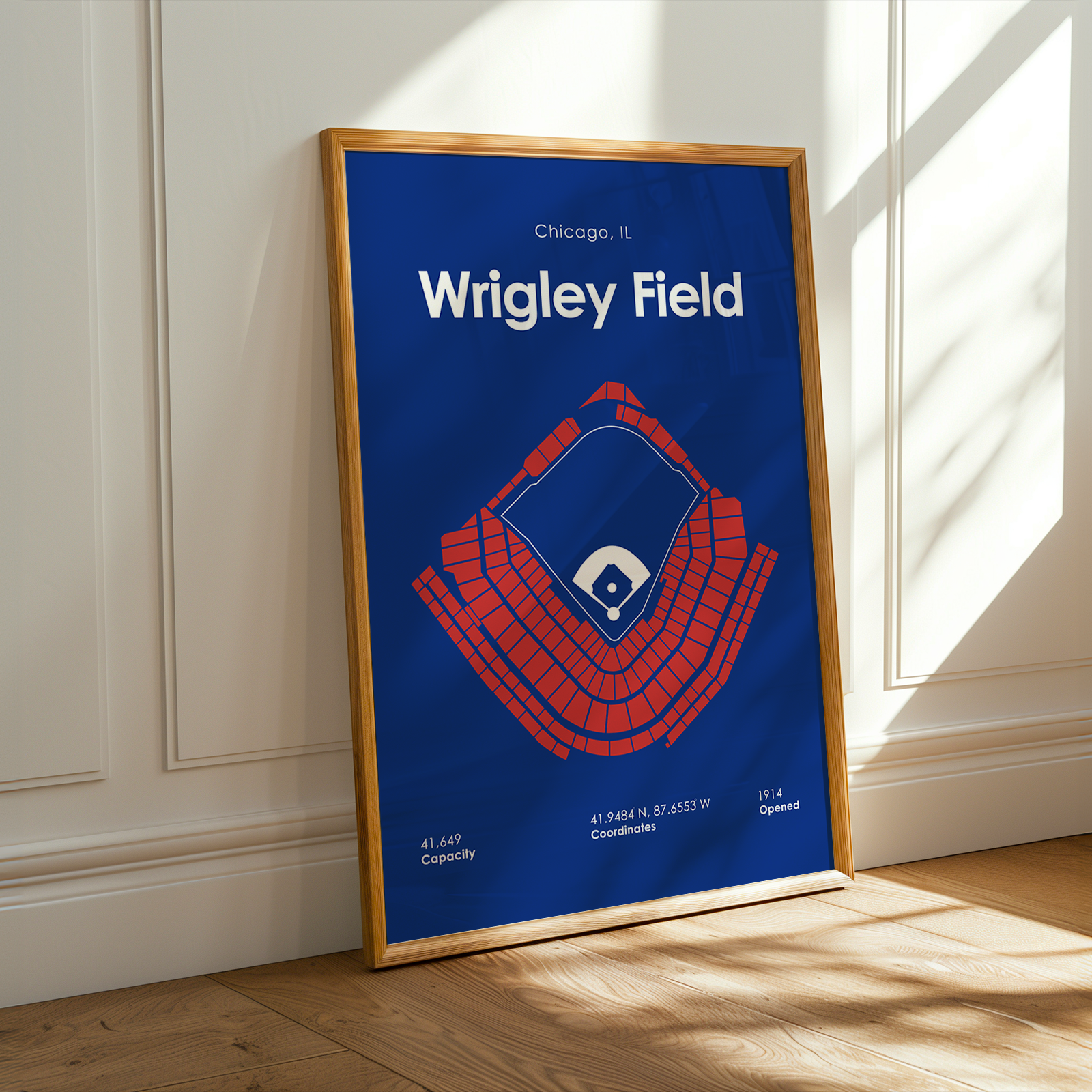 Framed poster of Wrigley Field with blue background and red stadium illustration, leaning against a white wall.