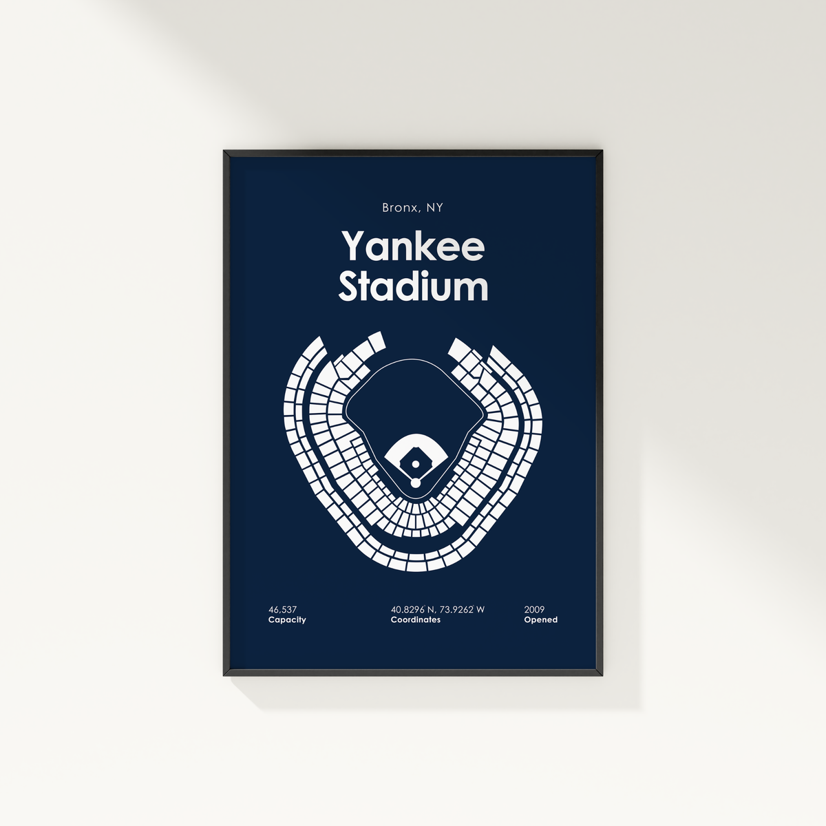 A framed minimalist poster of Yankee Stadium in the Bronx, NY, featuring a white stadium layout and text on a dark navy blue background, shown hanging on a light off-white wall.