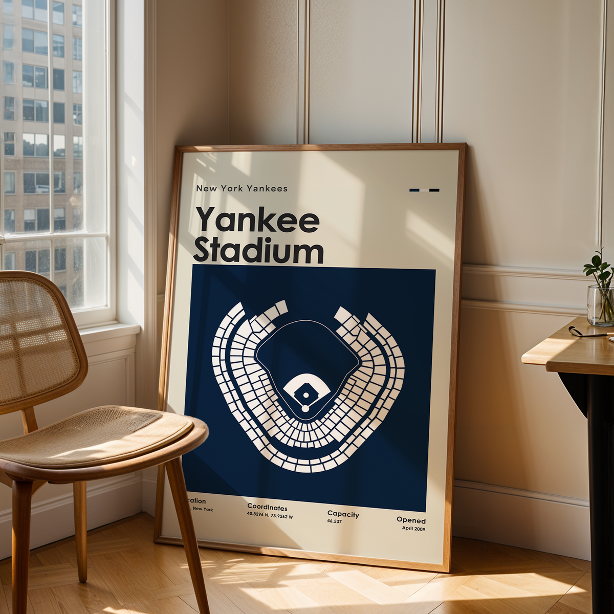A framed New York Yankees baseball stadium poster leans against a wall in a sunlit room with a wooden chair, table, and window.