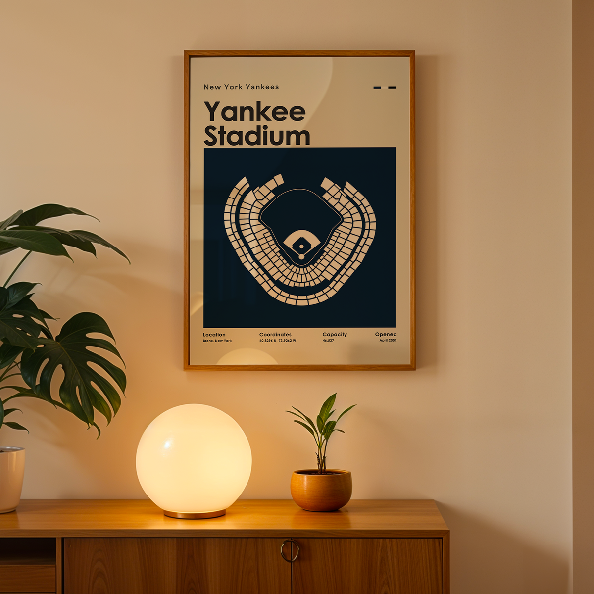 A framed Yankee Stadium poster hangs on a beige wall above a wooden console table. The poster features a cream-colored stadium seating diagram on a dark navy background.